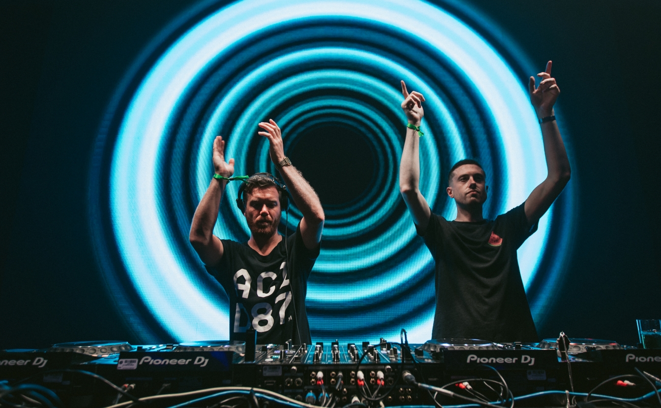 Two djs performing energetically at a club, with one arm raised each, standing in front of a mesmerizing circular blue light display and a table filled with dj equipment.