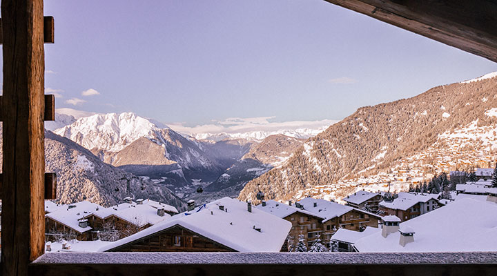 Snow-covered rooftops of a mountain village are seen from a wooden balcony, with tall, snowy peaks and a valley stretching into the distance under a clear sky.