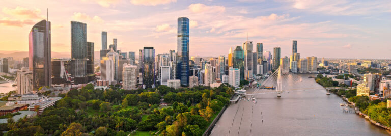 Vista panorámica del perfil de una ciudad moderna al atardecer, con altos rascacielos, un río que atraviesa la ciudad, un puente peatonal y exuberantes zonas verdes en primer plano.