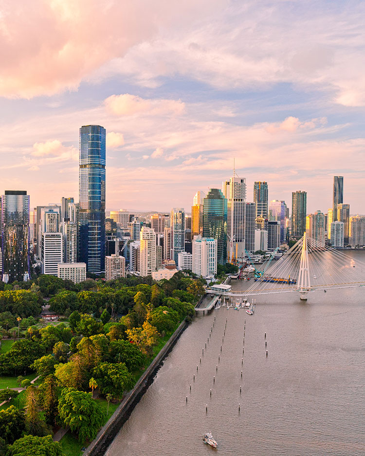 Aerial view of a modern city skyline with tall buildings, a river, a large green park, and a white cable-stayed bridge at sunset. A few boats are visible on the water.