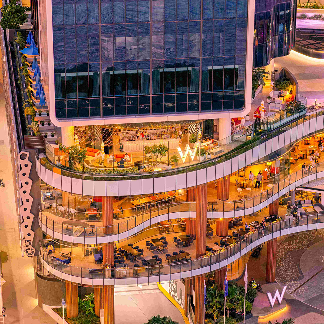 A modern, multi-story building with wavy terraces featuring outdoor seating areas, warm lighting, plants, and glass railings. People are dining and walking. Large lit 