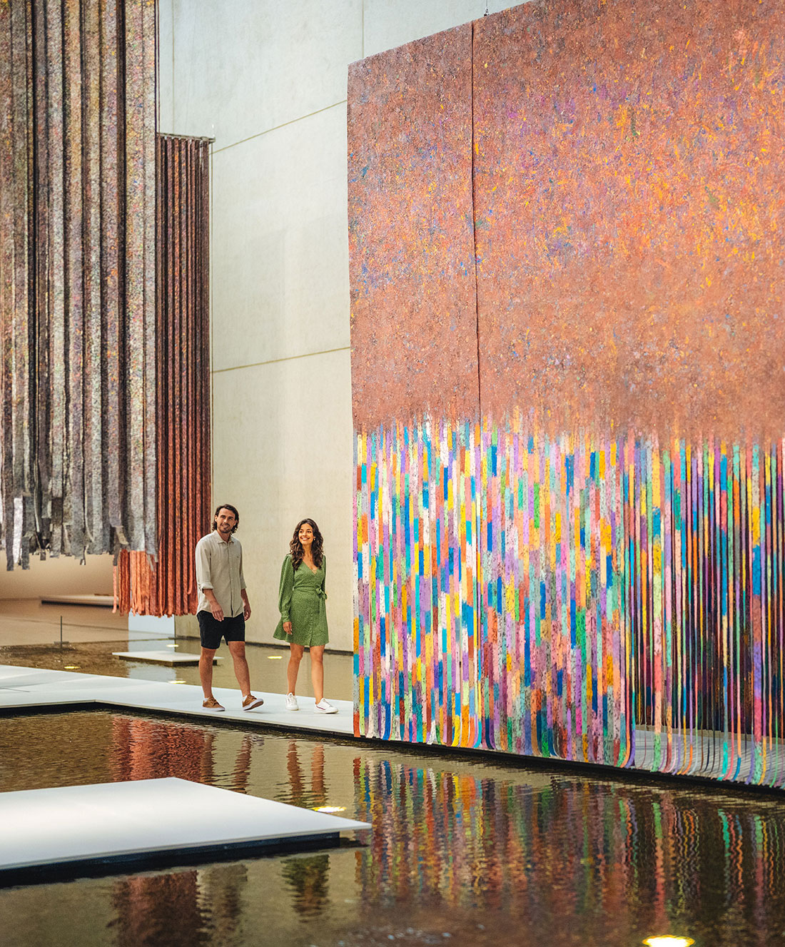 A man and a woman walk beside large, colorful abstract artworks and hanging fabric installations in a modern gallery with water features and white stepping platforms.