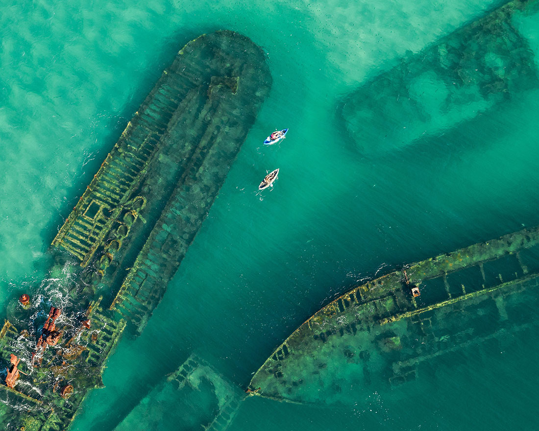 Aerial view of several sunken shipwrecks in clear turquoise water, with two small boats paddling between the rusted remains.