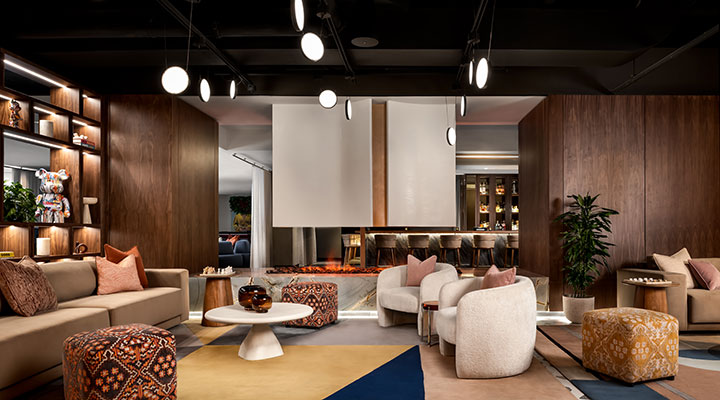 Modern lounge with neutral sofas, patterned ottomans, and armchairs arranged around a central fireplace. Warm wood-paneled walls, shelves with decorative items, and hanging lights create a cozy, stylish atmosphere.