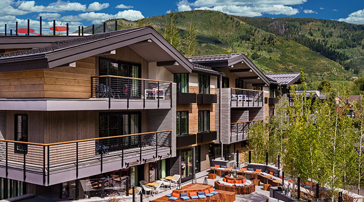 Modern mountain resort with wood and glass balconies, outdoor seating area with fire pits, and lush green hills and blue sky in the background.