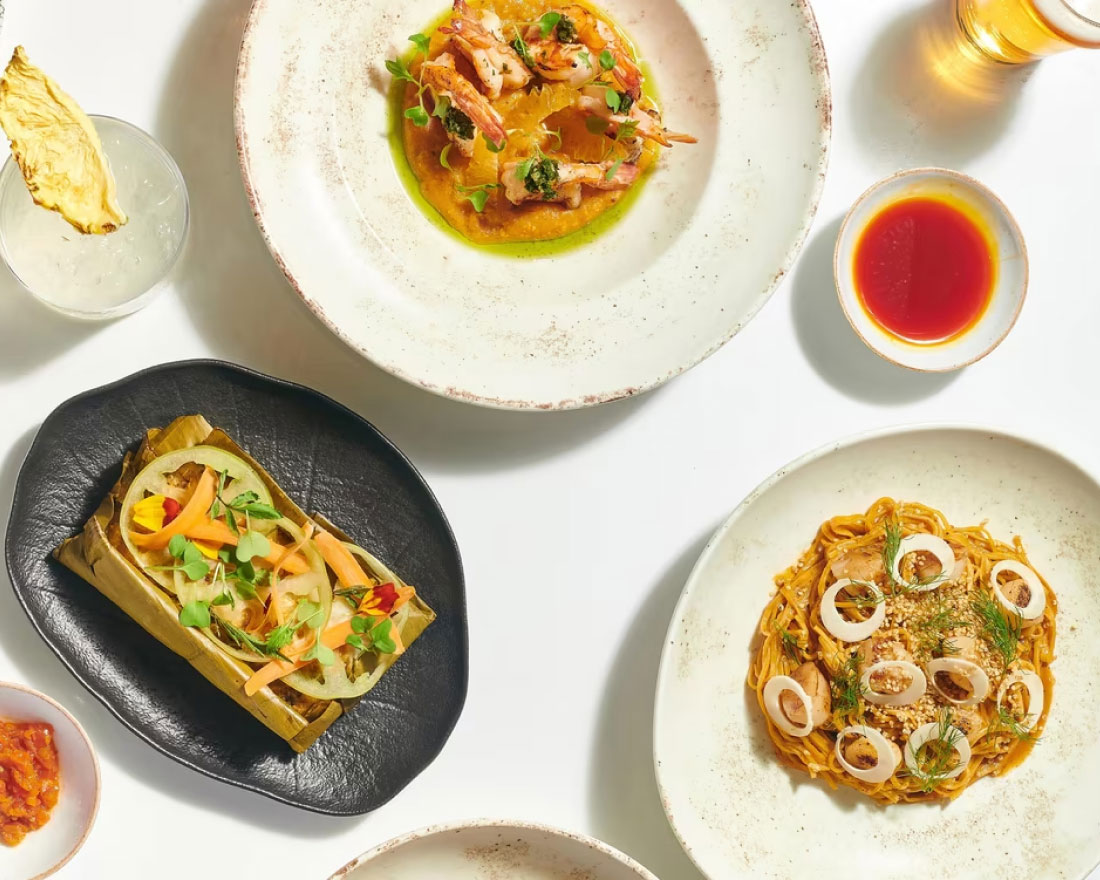 A table set with various dishes including a tamale wrapped in banana leaves on a black plate, a pasta dish garnished with herbs and sliced eggs on a white plate, a seafood dish with sauce, a small bowl of red sauce, a drink garnished with a pineapple slice, and a glass of beer.