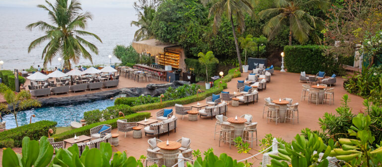 La terraza del restaurante al aire libre, con mesas y sillas, ofrece vistas a la piscina, a una exuberante vegetación y al mar. Hay sombrillas y una zona de bar cerca del agua, rodeada de palmeras y plantas tropicales.