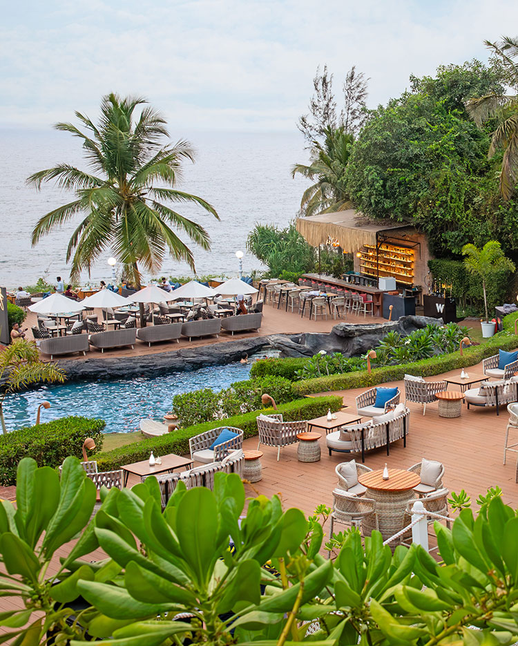 Outdoor resort with a pool, lounge chairs, umbrellas, bar, palm trees, and ocean view in the background. Green plants and seating areas surround the pool. Bright and tropical atmosphere.