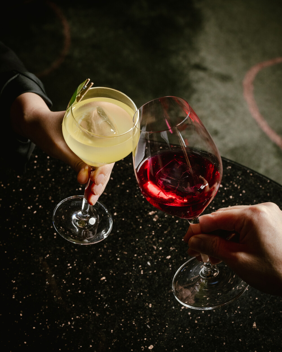 Two hands hold glasses of yellow and red cocktails while toasting over a dark, speckled table.