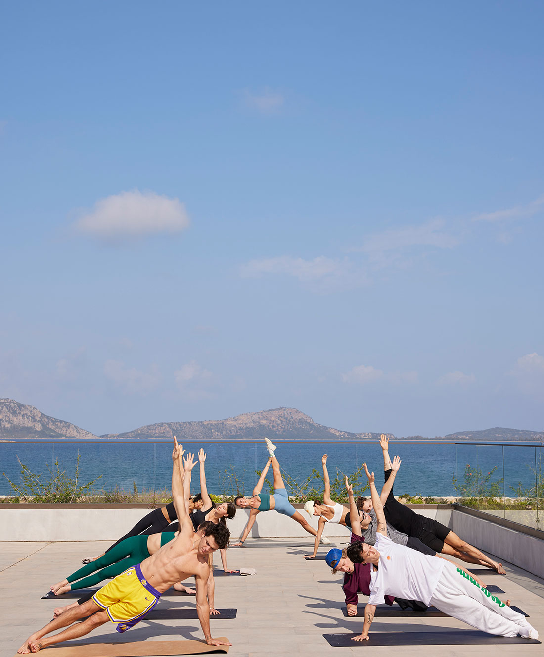 Eine Gruppe von Menschen praktiziert Yoga im Freien auf einer Terrasse am Meer. Sie führen seitliche Plankenhaltungen mit einem erhobenen Arm aus, unter einem klaren blauen Himmel mit fernen Bergen im Hintergrund.