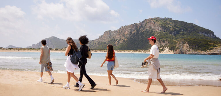 Cinco jóvenes caminan por una playa de arena cerca de aguas azules transparentes, con verdes colinas y acantilados rocosos de fondo bajo un cielo parcialmente nublado.