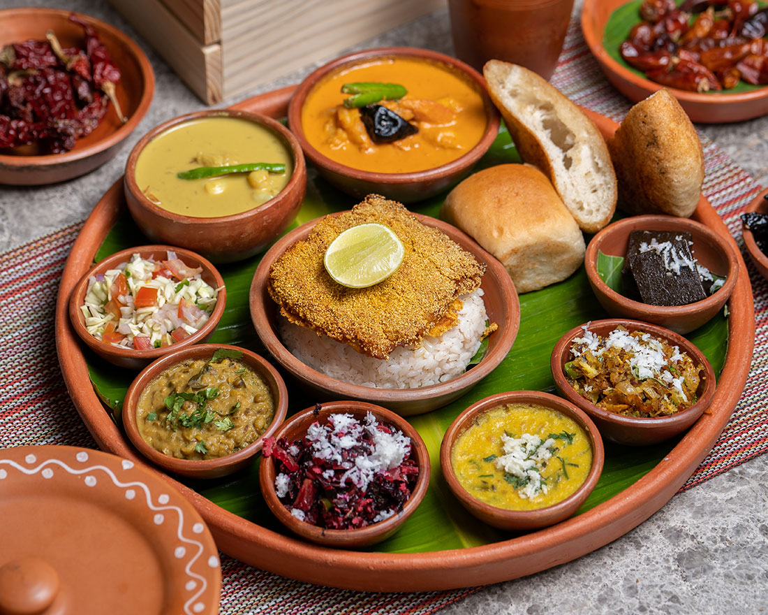 Un thali indio tradicional con varios platos en pequeños cuencos de barro, que incluyen curry, verduras, ensalada, lentejas, una chuleta frita con arroz, un panecillo y un postre, todo servido en un plato redondo con una hoja de plátano.