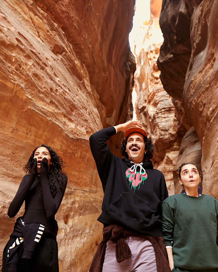 Three people stand between tall, rocky canyon walls, looking up in awe. Two appear excited or surprised, while the third stands calmly. Sunlight streams down from above, illuminating the scene.