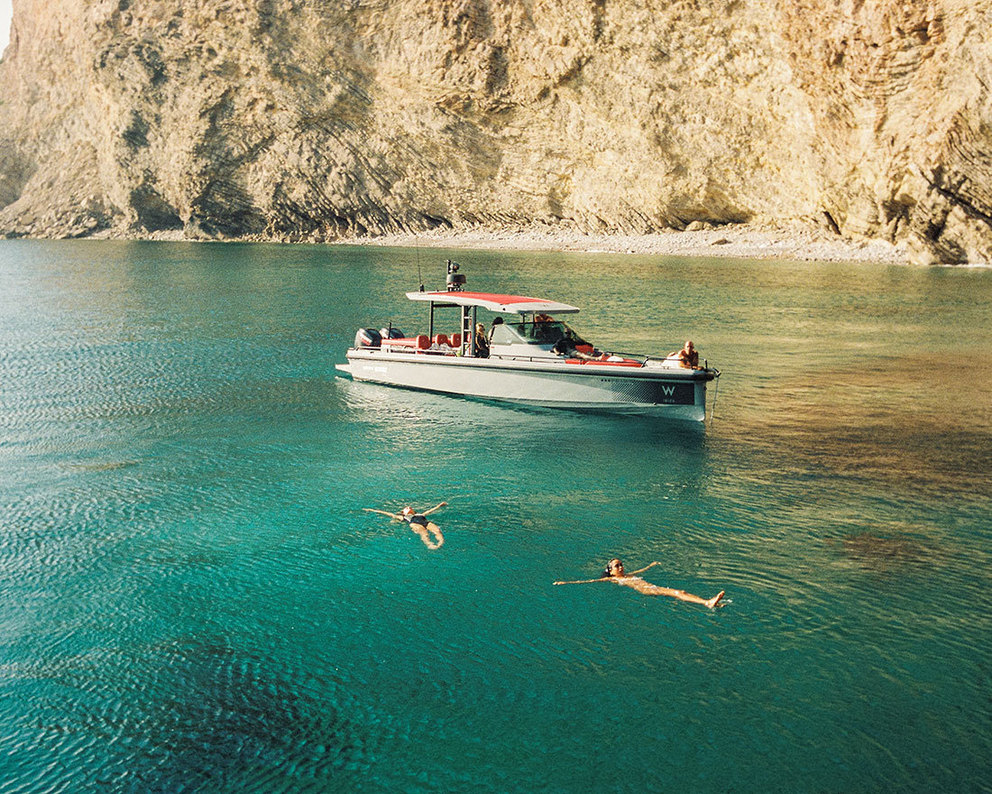 Una pequeña embarcación flota en aguas azules transparentes cerca de acantilados rocosos mientras dos personas nadan y flotan de espaldas en el mar cercano.