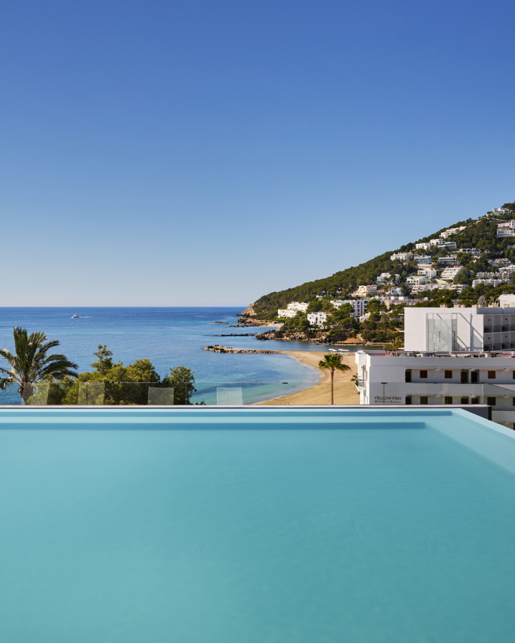 An infinity pool overlooking a sandy beach and coastal resort town under a clear blue sky.