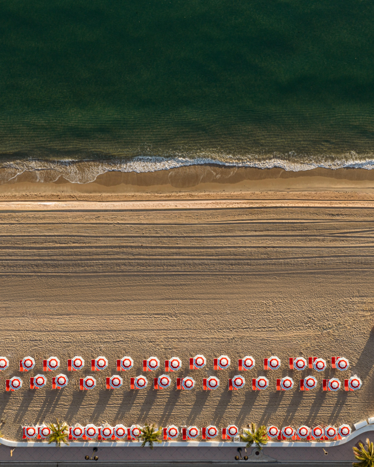 Vista aérea de una playa de arena con hileras ordenadas de sombrillas rojas y blancas, un mar verde y tranquilo, y olas suaves que se acercan a la orilla.