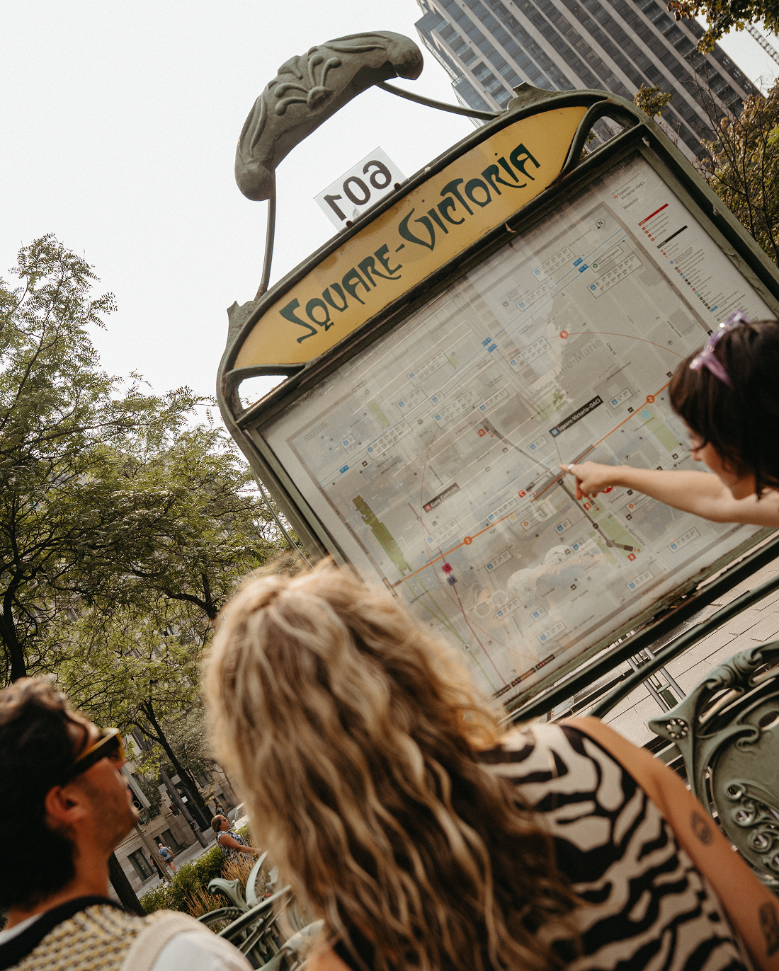 Tres personas miran un mapa grande en la plaza Victoria; una de ellas señala un lugar, con árboles y edificios altos al fondo.