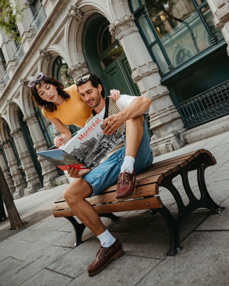 Una pareja sonriente está sentada en un banco frente a un edificio histórico, mirando juntos un gran mapa de la ciudad de Montreal. La mujer se asoma por encima del hombro del hombre mientras planifican su recorrido turístico.