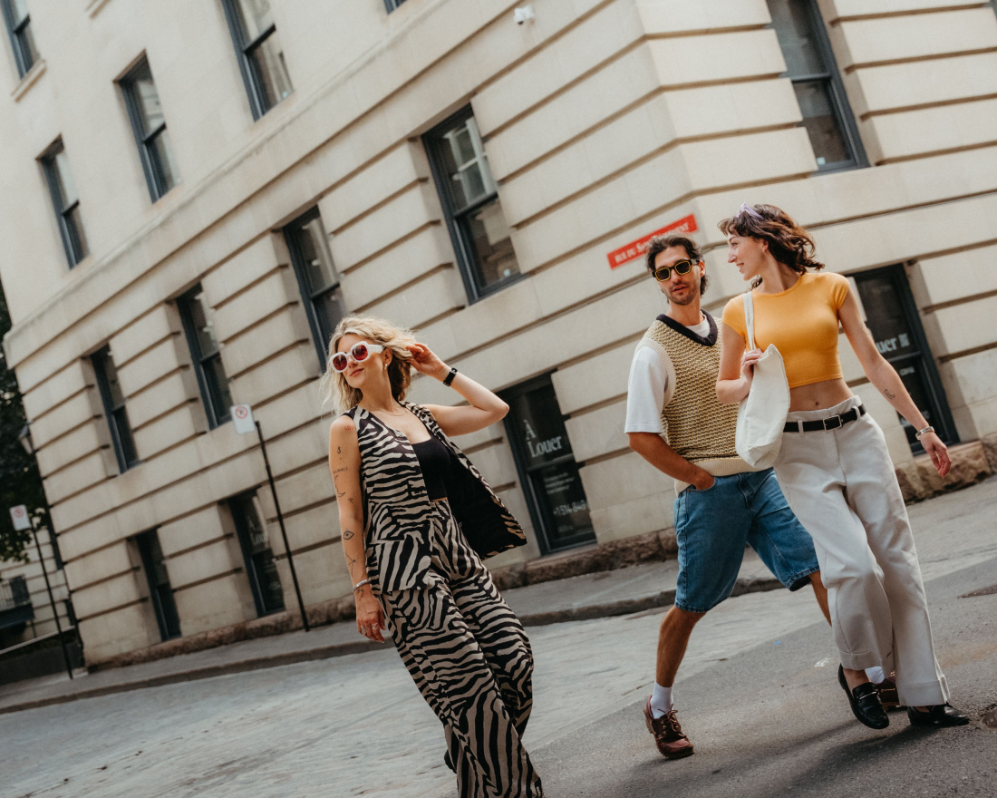 Tres personas caminan por una calle de la ciudad junto a un edificio de color beige; una de ellas, vestida con un conjunto con estampado de cebra, va delante, mientras que las otras dos, con ropa informal de verano, caminan detrás, en medio de una conversación.