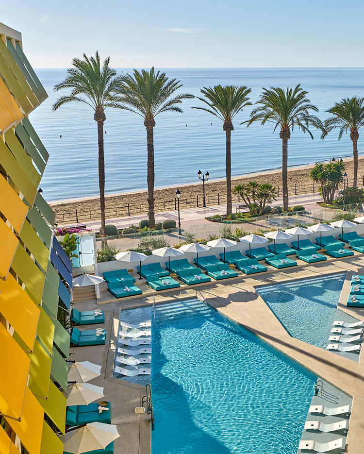 A beachfront resort features a pool area with teal lounge chairs and umbrellas, tall palm trees, and a sandy beach leading to calm blue ocean under a clear sky. A modern yellow building is visible on the left.