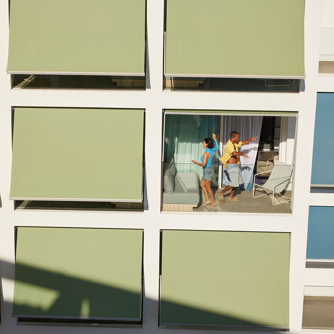 Vista de la fachada de un edificio con varios toldos verdes cubriendo las ventanas. En una ventana abierta, dos personas están en un balcón, hablando y haciéndose gestos. Las ventanas de alrededor están cubiertas.