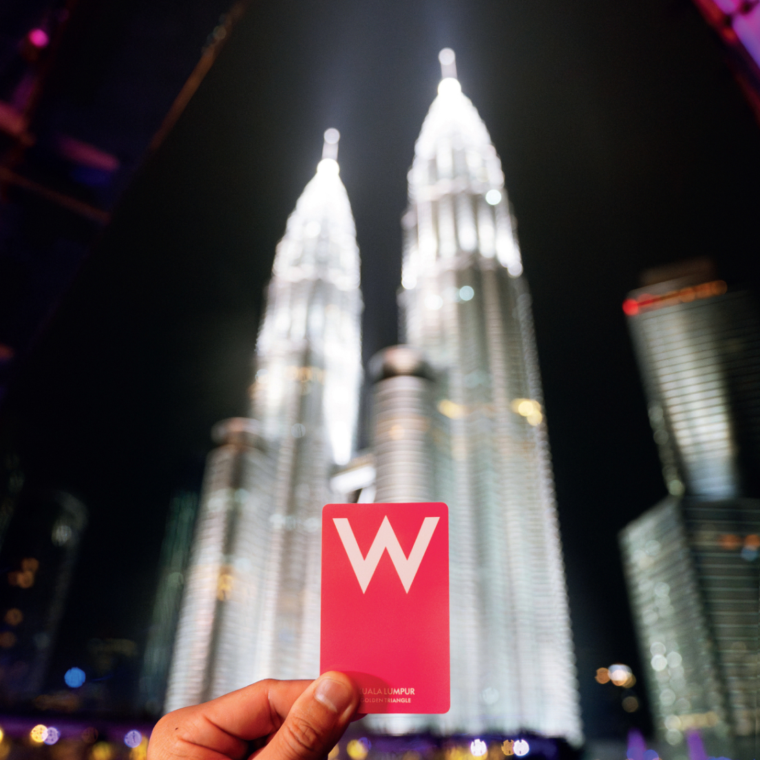 Una mano sostiene una tarjeta rosa con una «W» blanca delante de las Torres Gemelas Petronas iluminadas en Kuala Lumpur por la noche.