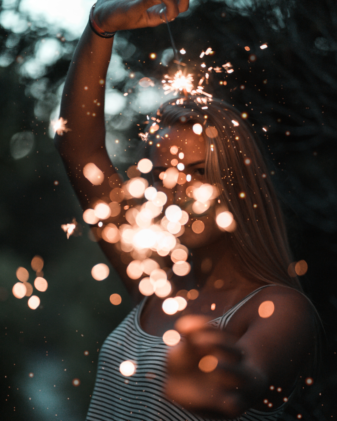 Una persona con una camiseta sin mangas a rayas sostiene unas bengalas, y las chispas brillantes y los efectos de bokeh le ocultan parcialmente el rostro. El fondo está desenfocado y oscuro, lo que resalta la luz de las bengalas.