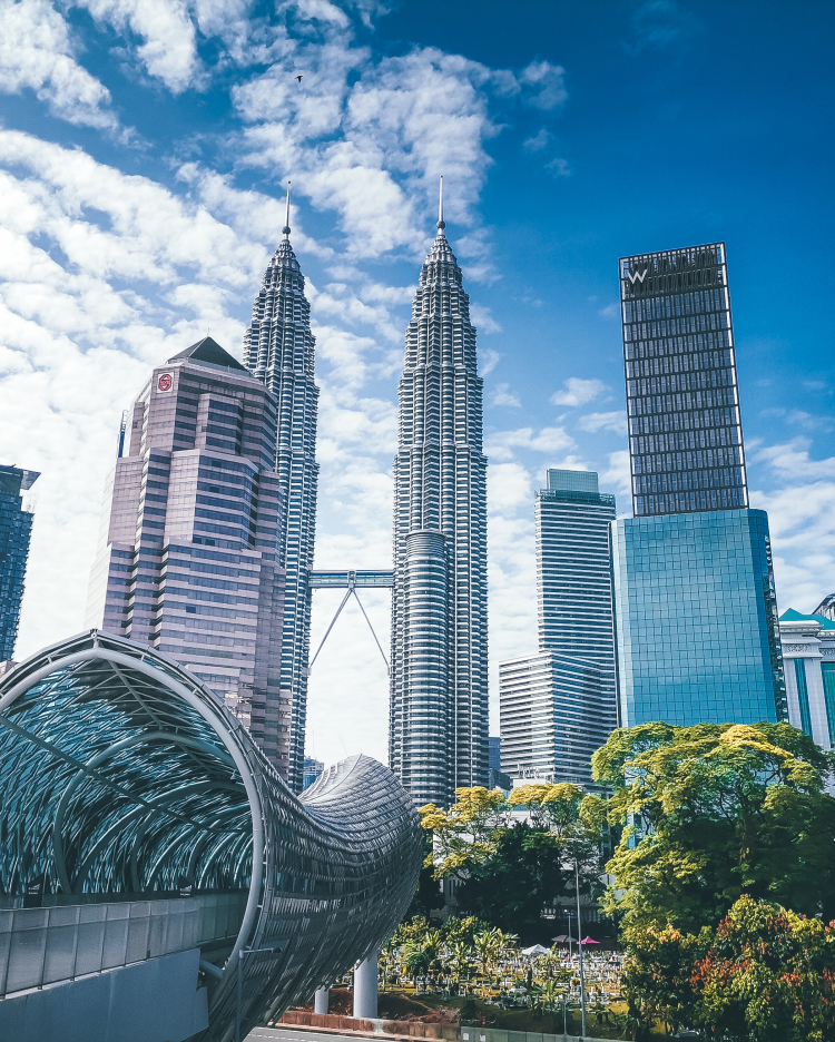 El perfil urbano de Kuala Lumpur, con las Torres Gemelas Petronas, los rascacielos circundantes, un moderno puente peatonal en primer plano y un cielo azul parcialmente nublado en lo alto.