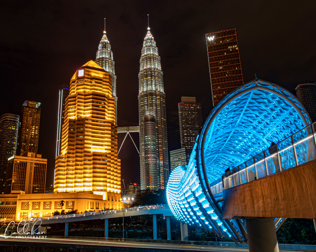 Vista nocturna de Kuala Lumpur con las Torres Gemelas Petronas iluminadas, un edificio dorado y un moderno puente peatonal que brilla en azul en primer plano.