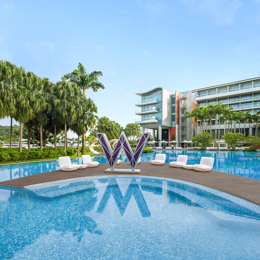 A modern hotel with glass balconies overlooks a large outdoor pool featuring a purple 