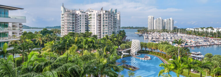 Una zona residencial tropical frente al mar con altos y modernos condominios, una gran piscina, palmeras y un puerto deportivo lleno de barcos bajo un cielo azul.
