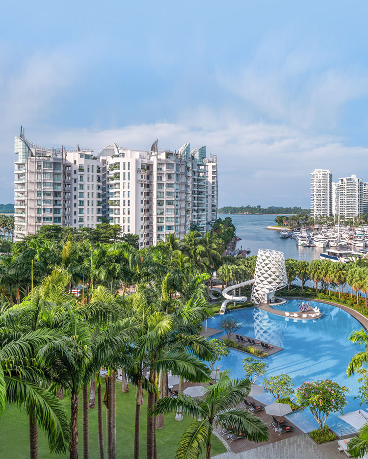 A luxury resort featuring tall white buildings, a large blue swimming pool, palm trees, loungers, and a marina with boats docked along the waterfront under a partly cloudy sky.