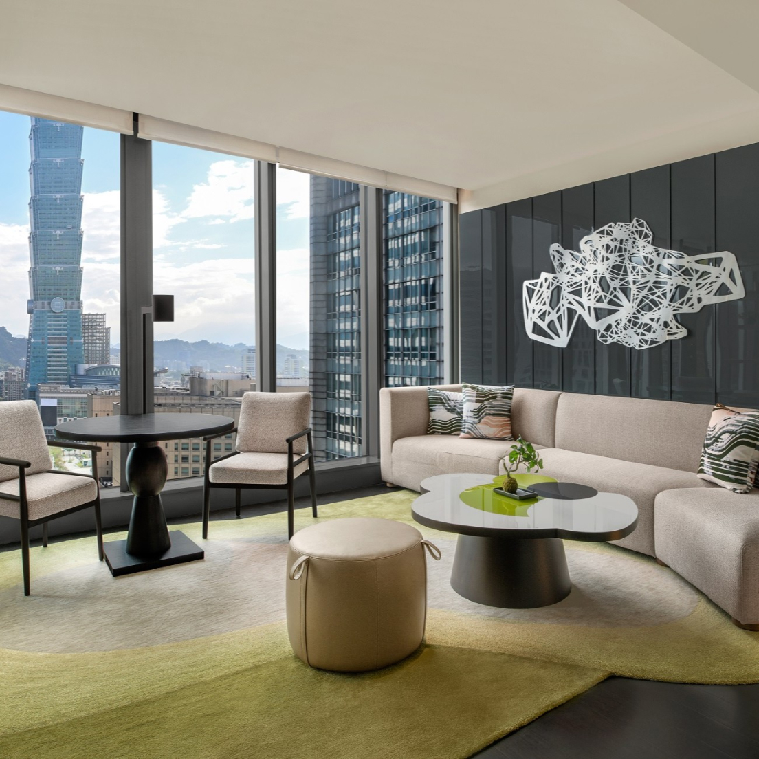A modern hotel lounge with a sofa, circular coffee table, and small dining set, featuring a view of Taipei 101 through floor-to-ceiling windows.