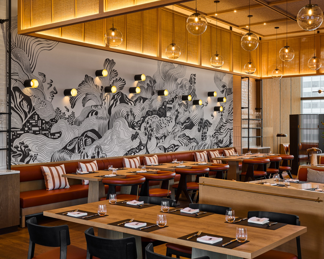 A dining room with wooden tables, black chairs, and a long banquette against a wall featuring a large, intricate black-and-white nature-inspired mural.