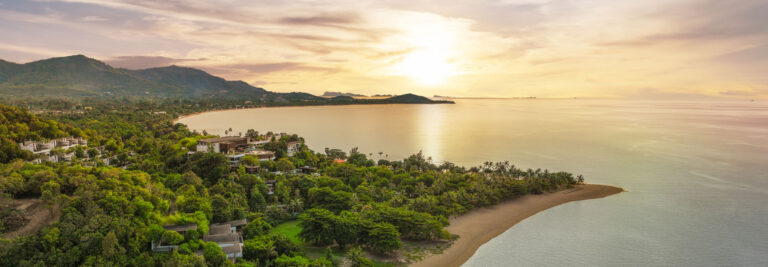 Vista aérea de una exuberante costa verde con edificios, playa de arena y mar en calma bajo un cielo dorado al atardecer, con montañas al fondo.