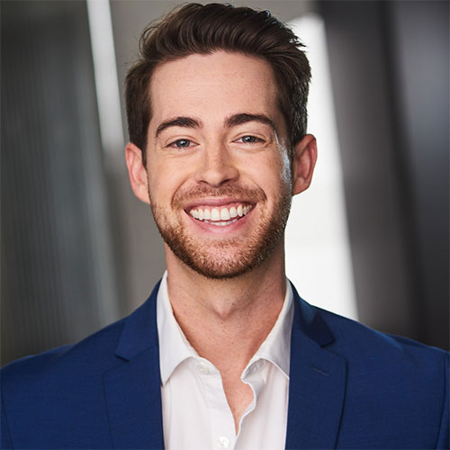 A smiling man with short brown hair and a trimmed beard, dressed in a blue suit jacket and a white shirt, stands in front of a blurred background. He looks directly at the camera.