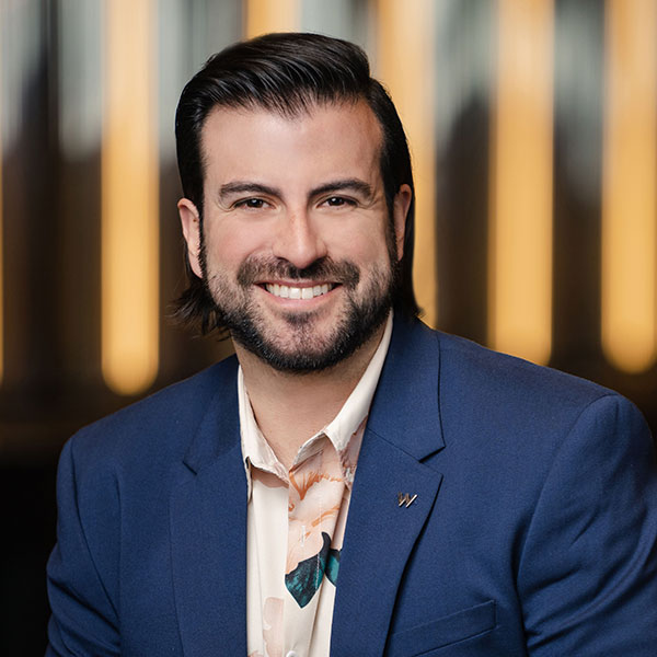 A smiling man with dark hair and a beard is wearing a blue blazer over a light-colored shirt with a floral pattern. He is positioned in front of a warmly lit background with vertical light strips.