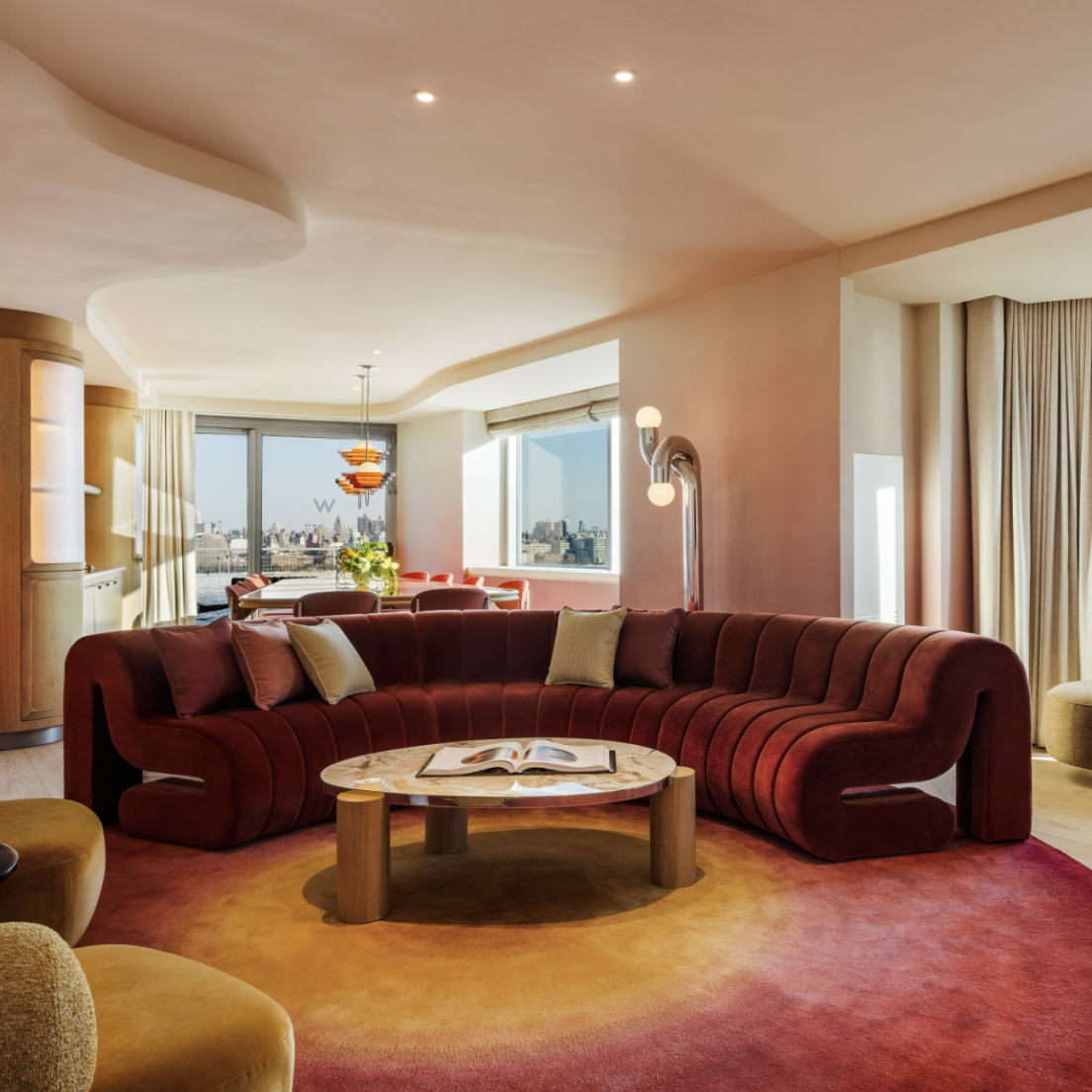 A curved, burgundy velvet sofa sits on a circular gradient rug in front of a marble coffee table, with a city skyline visible through large windows in the background.