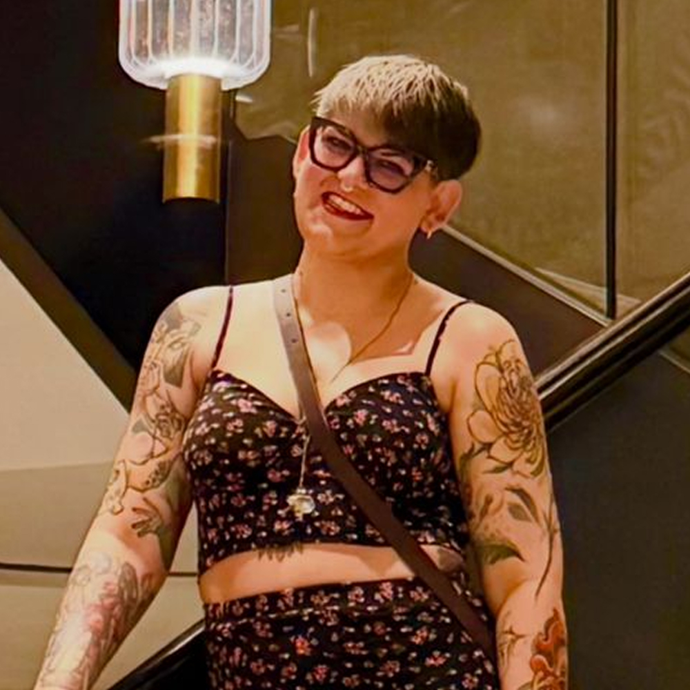 Una persona con el pelo corto, gafas y tatuajes florales lleva un top corto negro con motivos florales y sonríe, de pie en el interior, cerca de una escalera y de un aplique moderno.