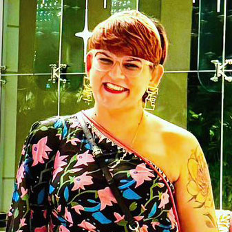 A person with short brown hair and glasses is smiling. They are wearing a one-shoulder top with a floral pattern and have visible tattoos on their arm. The background includes a reflective surface with geometric designs.