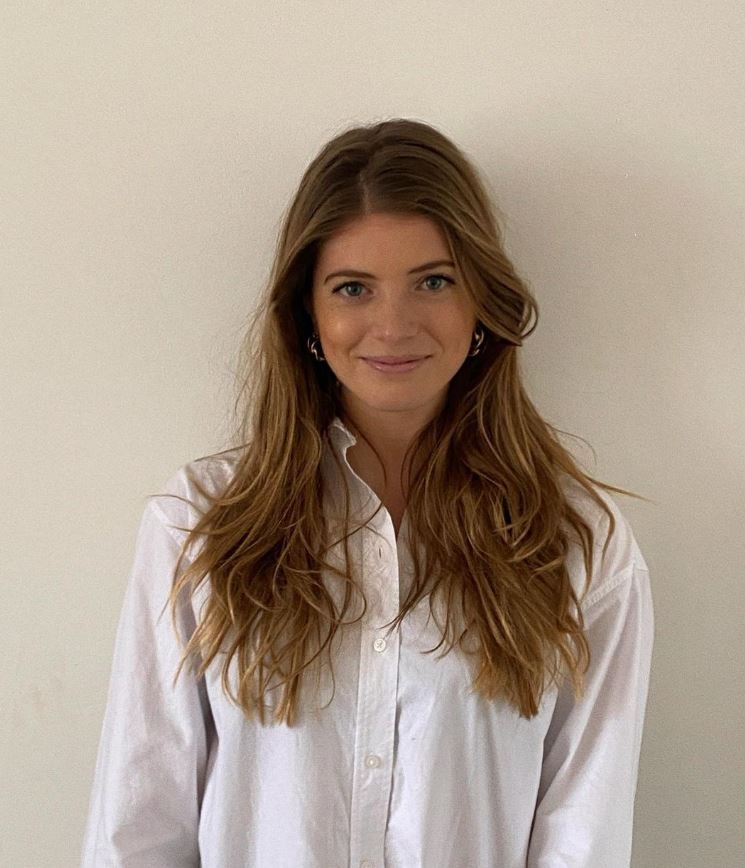 A woman with long, wavy brown hair wearing a white shirt smiles subtly while standing against a plain beige wall.