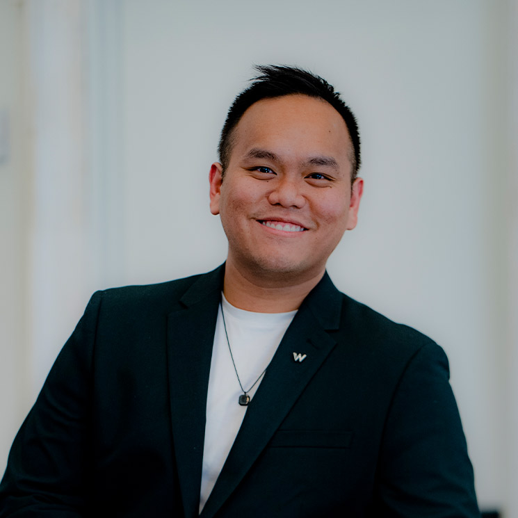 A smiling asian man wearing a black blazer and a white shirt, sitting in a well-lit room with a blurred white background. he has short black hair and a necklace with a pendant.