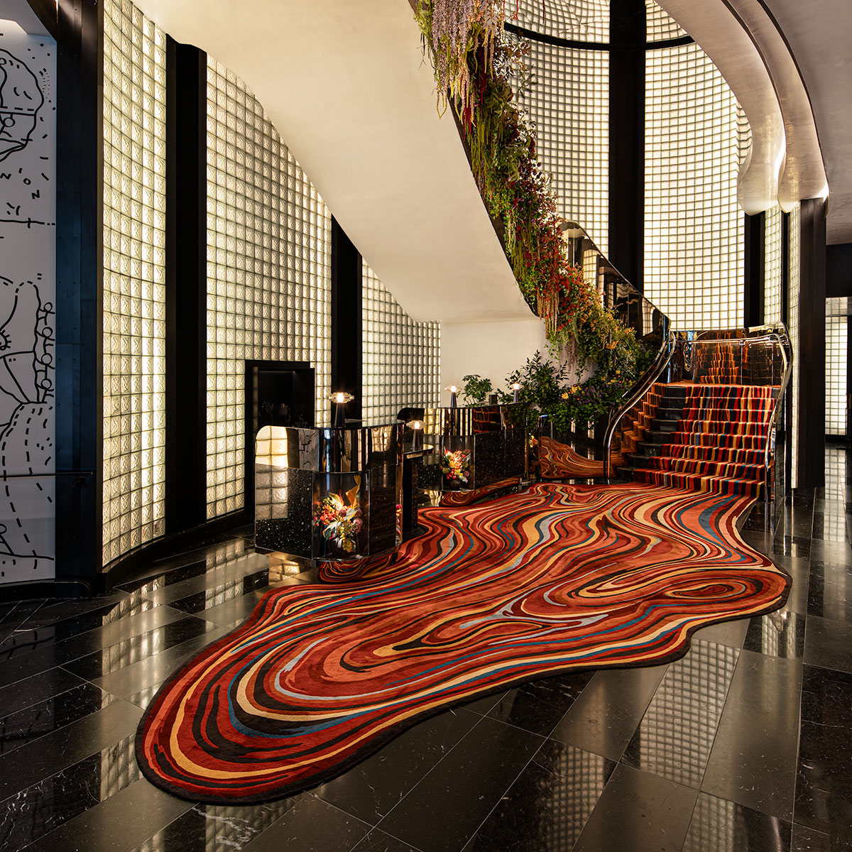 A modern, luxurious lobby with a black marble floor, a colorful wavy-patterned carpet, a grand staircase adorned with flowers, and walls of glass blocks. Warm lighting creates an elegant and inviting atmosphere.