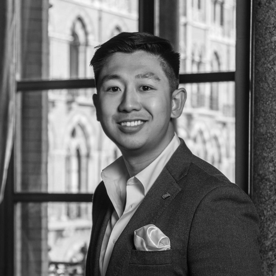 A black and white portrait of a smiling asian man wearing a suit, with a blurred architectural background visible through a window.