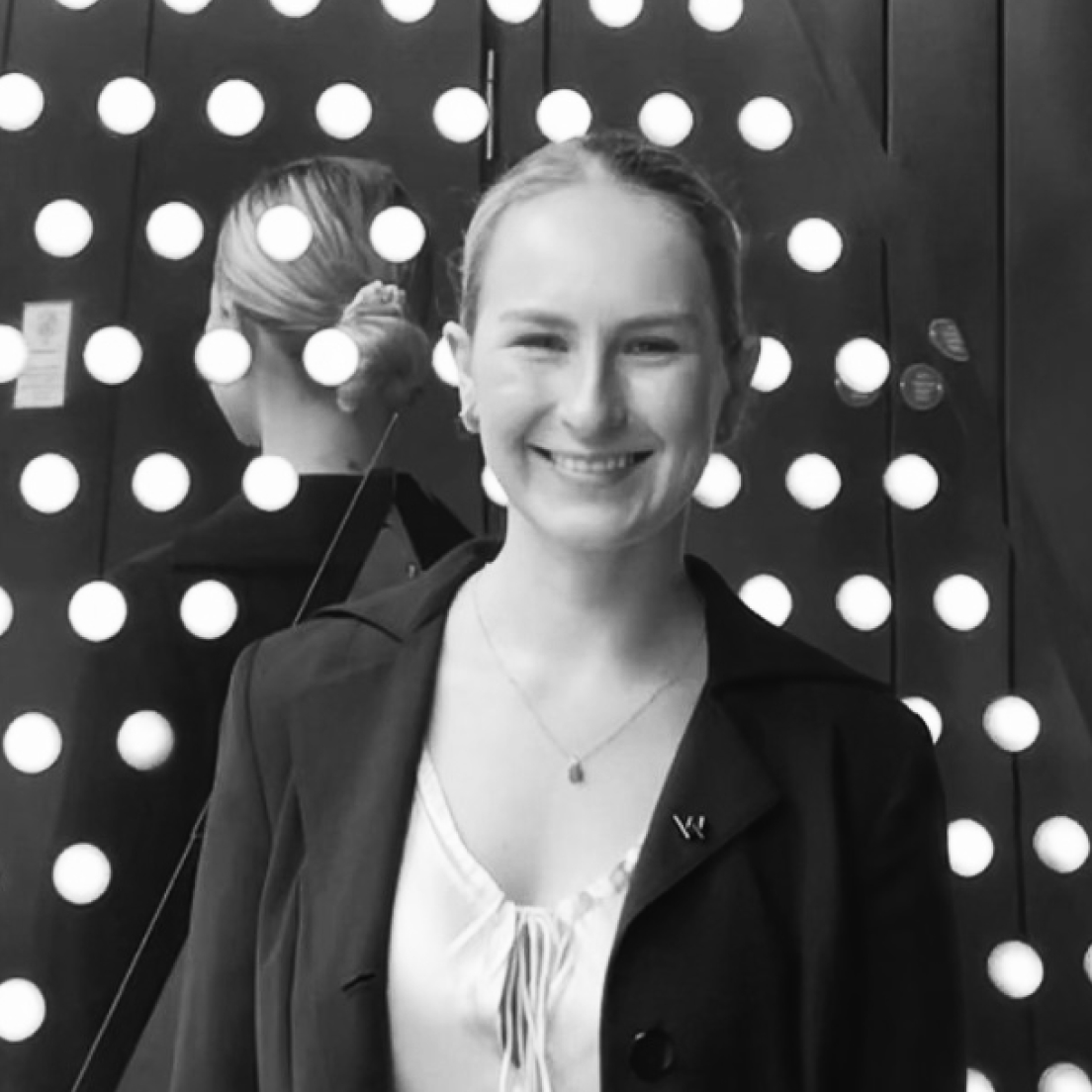Black and white portrait of a smiling young woman with her hair styled in a bun, wearing a blazer and blouse. she stands in front of a wall adorned with circular lights.