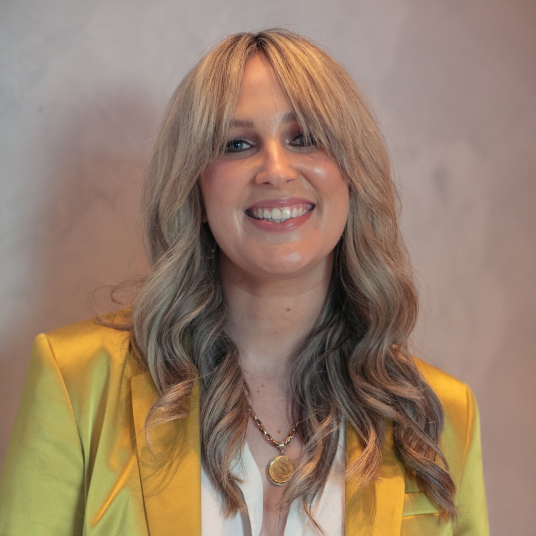 A smiling woman with medium-length wavy blonde hair, wearing a yellow blazer and a gold necklace, standing in front of a textured pink background.