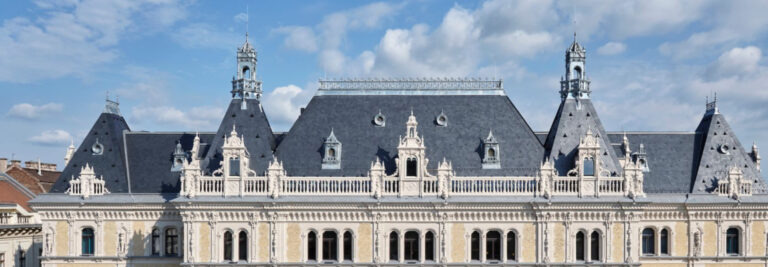 A panoramic view of a grand historical building with ornate architecture, featuring multiple spires, intricate carvings, and a symmetrical façade under a clear blue sky.