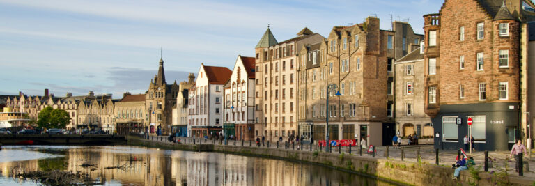 Historic buildings line the sunlit banks of a calm river reflecting the clear blue sky in a tranquil urban scene, with pedestrians walking along the riverfront in Edinburgh.