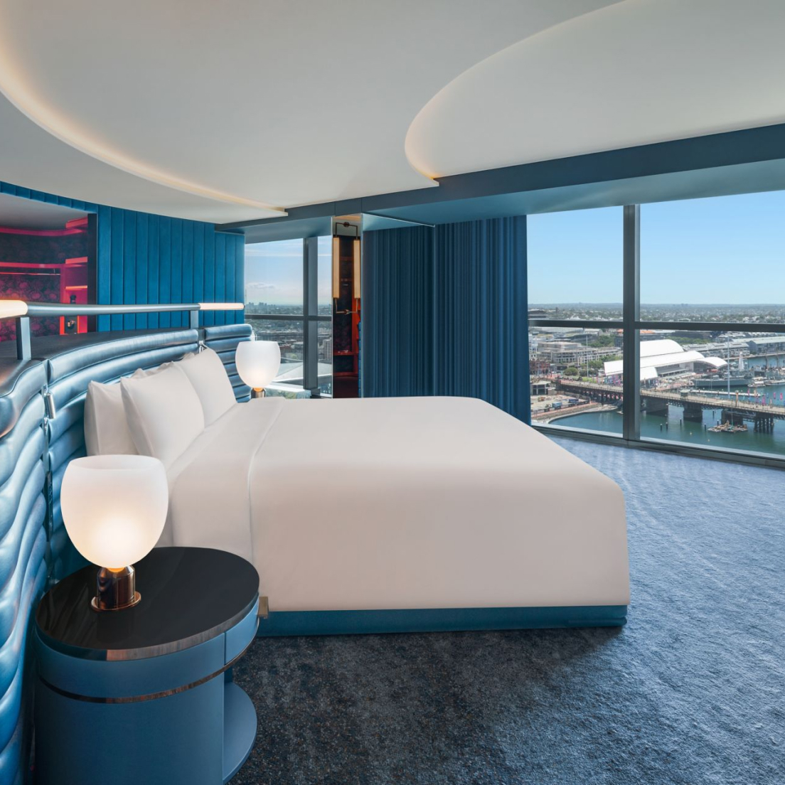 Luxurious hotel room featuring a large bed with white bedding, blue tufted headboard, and blue curtains, overlooking a cityscape through floor-to-ceiling windows.