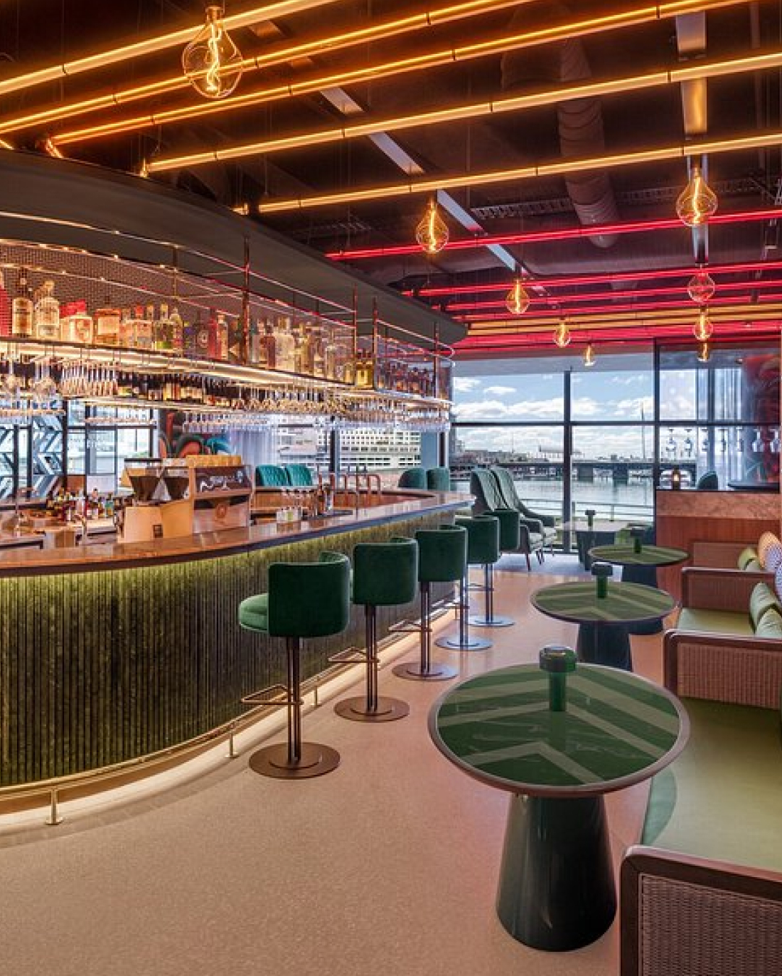 A stylish bar interior with green velvet stools, a curvaceous green counter, and an array of liquor bottles. the ceiling has distinctive red glow lighting, and large windows offer a bright view.